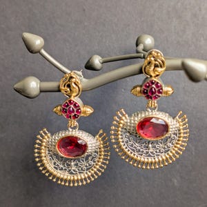 May include: A pair of ornate earrings with a silver and gold design. Each earring features a large, oval, red gemstone at the center, surrounded by intricate metalwork. The earrings are displayed on a gray jewelry stand.