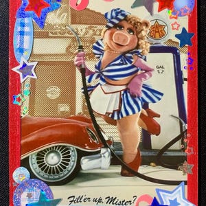 May include: A trading card featuring Miss Piggy in a blue and white striped outfit, holding a gas pump. A red classic car and gas pump are in the background. The card has decorative stars and the text "Filler up, Mister?"