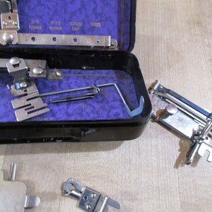 May include: A set of vintage sewing machine attachments in a black metal case with a purple velvet lining. The attachments include a variety of feet, needles, and other tools. The case is open and the attachments are visible.
