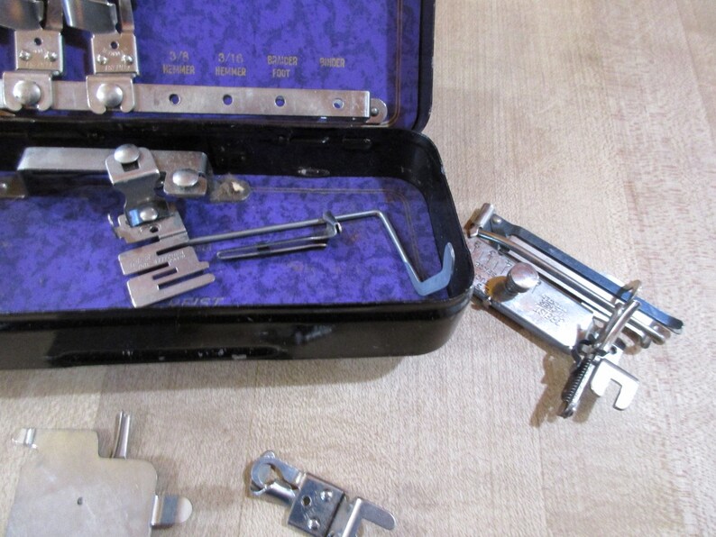 May include: A set of vintage sewing machine attachments in a black metal case with a purple velvet lining. The attachments include a variety of feet, needles, and other tools. The case is open and the attachments are visible.