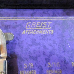 May include: Close-up of a purple sewing machine case with gold lettering that reads "Greist Attachments". The case also has markings for "3/8 Hemmer" and "3/16 Hemmer" and "Braider Foot".