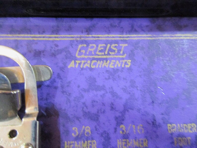 May include: Close-up of a purple sewing machine case with gold lettering that reads "Greist Attachments". The case also has markings for "3/8 Hemmer" and "3/16 Hemmer" and "Braider Foot".