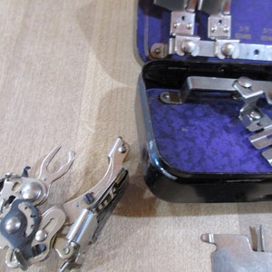 May include: A silver sewing machine foot attachment with a spring-loaded mechanism. The foot is in a raised position and appears to be made of metal. The foot is part of a sewing machine accessory kit.