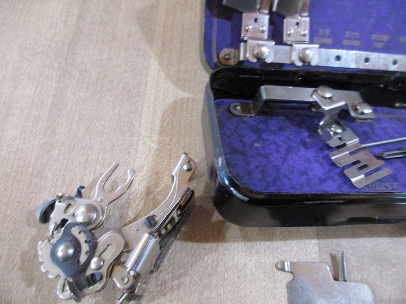 May include: A silver sewing machine foot attachment with a spring-loaded mechanism. The foot is in a raised position and appears to be made of metal. The foot is part of a sewing machine accessory kit.