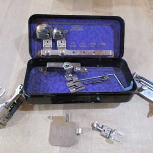 May include: A set of vintage sewing machine attachments in a black metal case with a purple velvet lining. The case is open and the attachments are visible. The attachments include a variety of feet, needles, and other tools. The case has the text "GRIST ATTACHMENTS" printed on the front.