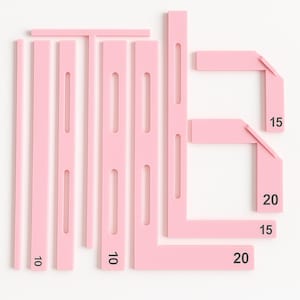 May include: A set of pink plastic drafting tools, including rulers and right-angled triangles. The tools are marked with black numbers, including 10, 15, and 20. The tools are arranged on a white surface.