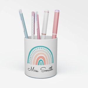 May include: White ceramic pencil holder with a rainbow design and the text "Mrs. Smith". The holder has five pens inside, including pink, blue, and white.