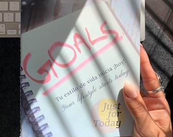 May include: A white notebook with a pink inscription "GOALS" and the text "Just for Today" in yellow and orange. The notebook has a spiral binding and the text "Tu estilo de vida inicia ¡hoy! Your lifestyle starts today" is printed on the cover.