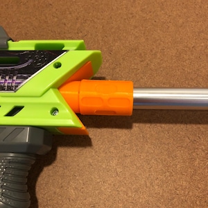 May include: A close-up of a toy gun with a green and orange plastic design. The gun has a silver barrel and a gray handle. The green plastic has a decorative design.