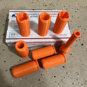 May include: A collection of seven bright orange plastic cylindrical adapters for toy foam dart blasters. Each piece features a hollow center and embossed text on the side, including the words RIVAL, ELITE, and MEGA, with varying internal structures.