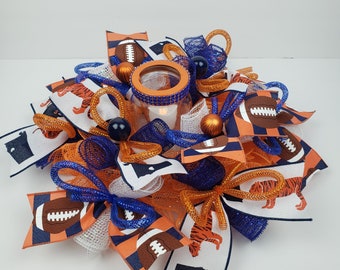 May include: A football-themed centerpiece with orange, blue, and white mesh, ribbons, and fabric. The centerpiece features a clear glass jar with a teal blue rim and a white candle inside. The fabric features a football design and a tiger graphic.