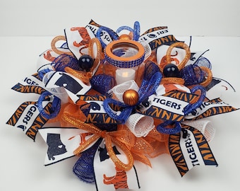 May include: Orange and blue mesh wreath with a clear glass votive holder in the center. The wreath is decorated with ribbons that say "Tigers" and have a tiger print. There are also black and white silhouettes of a tiger head and a state outline.