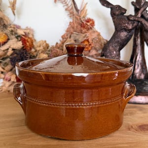 May include: A round, brown glazed ceramic casserole dish with a matching lid. The pot features two small side handles and a decorative beaded texture band encircling the middle. The lid has a central knob handle and a smooth, glossy finish.