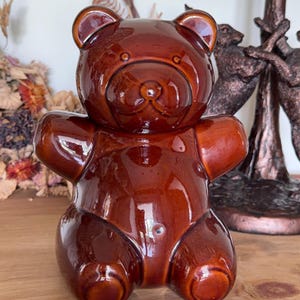 May include: A glossy, dark brown ceramic bear figurine in a seated position. The bear has a smooth, reflective surface, rounded ears, a defined snout, and a simple, moulded body shape with outstretched arms and visible feet, creating a solid form.