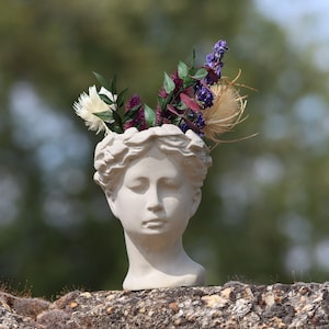 Könnte beinhalten: Eine weiße Gips-Kopfskulptur einer Frau, die als Vase dient und mit lila, weißen und grünen Blumen gefüllt ist. Die Skulptur steht auf einer verwitterten Steinmauer, mit einem verschwommenen grünen Hintergrund. Der Kopf ist ein klassisches Design.