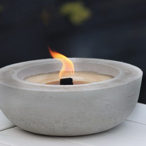 May include: A round, light grey concrete bowl containing a lit candle with a dark wick. The candle wax is a pale yellow colour, and a small, flickering orange and yellow flame rises from the centre of the bowl, casting a warm glow.