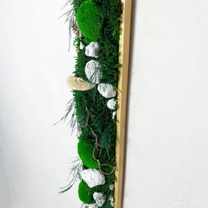 Könnte beinhalten: Ein vertikaler Wandbehang mit einem Holzrahmen und einem grünen Moosdesign. Das Moos ist in einem kaskadierenden Muster mit weißen Steinen und kleinen Treibholzstücken angeordnet.