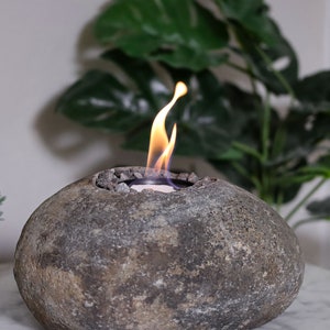 Könnte beinhalten: Ein grauer Stein-Feuerstelle mit einer kleinen Flamme, die darin brennt. Die Feuerstelle ist in Form eines großen, runden Steins geformt und hat eine kleine Öffnung oben für die Flamme. Die Feuerstelle steht auf einer weißen Oberfläche.