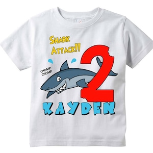 May include: White t-shirt with a cartoon shark graphic. The design includes the words "Shark Attack!!" in yellow, a large red number "2", and the name "Kayden" in blue. The shark is gray and white with a toothy grin.