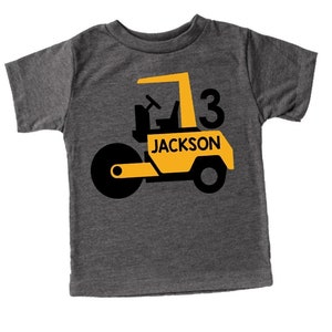 May include: A dark gray t-shirt featuring a yellow and black road roller design. The design includes the number 3 and the name "JACKSON" in a bold font. The shirt is a short-sleeved crew neck.