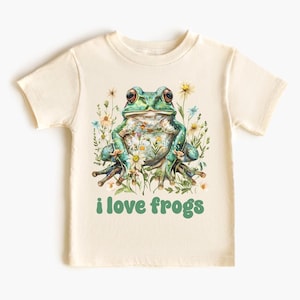 May include: A light beige t-shirt with a green frog sitting in a bed of flowers. The frog is surrounded by green leaves and white flowers. The text "i love frogs" is printed in green below the frog.