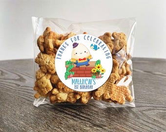 May include: A round sticker with a blue border and the text "Thanks for celebrating Matthew's 1st Birthday". The sticker features a cartoon illustration of an egg with a face, wearing a blue and white outfit, standing on a brick wall with flowers and a bird.