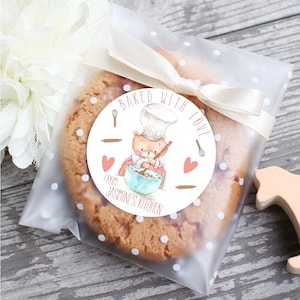 May include: A white round sticker with a brown bear wearing a chef's hat and holding a wooden spoon. The bear is mixing batter in a blue bowl. The sticker says "Baked with love from Jasmine's Kitchen".