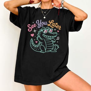 May include: Black t-shirt featuring a cartoon alligator wearing sunglasses, with the text "See You Later." The design includes hearts and stars. The shirt has a relaxed, oversized fit, suitable for casual wear.