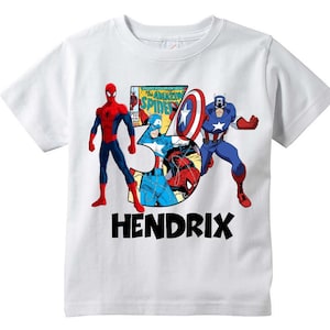 May include: White t-shirt with a red and blue superhero graphic featuring Spider-Man and Captain America. The graphic includes a comic book cover with the text "The Amazing Spider-Man". The shirt also has the name "HENDRIX" printed in black below the graphic.