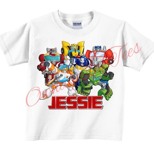 May include: White t-shirt with a colorful graphic of Transformers characters. The text "JESSIE" is printed in red below the characters.
