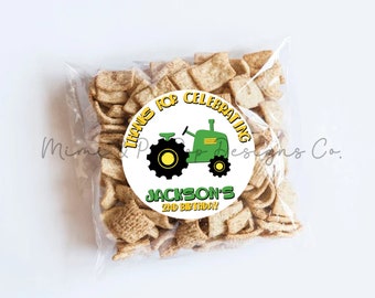 May include: A clear cellophane bag filled with square cereal pieces. A round sticker on the bag reads "THANKS FOR CELEBRATING" with a green tractor graphic and "JACKSON'S 2ND BIRTHDAY" text. The bag is on a white background.