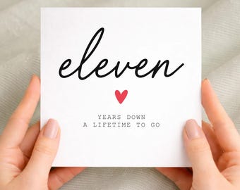 May include: A square white greeting card featuring the black cursive text eleven, a small red heart icon, and the black typewriter-style text YEARS DOWN A LIFETIME TO GO printed on the front surface of the card.
