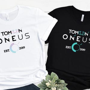 May include: Two t-shirts, one white and one black, with the text "TOMOON ONEUS" and "EST. 2019" printed on the front. The design includes a stylized moon and a circular graphic in teal and pastel colors. The shirts are short-sleeved.