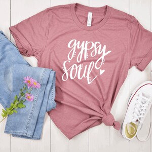 Puede incluir: Una camiseta color malva con el texto "gypsy soul" en letras blancas con un peque&ntilde;o coraz&oacute;n rosa. La camiseta se combina con unos vaqueros azules y zapatillas blancas.