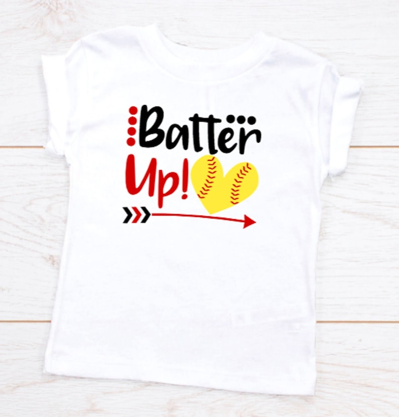May include: White t-shirt with black text that says "Batter Up!" with a red arrow pointing to the right and a yellow softball in a heart shape.