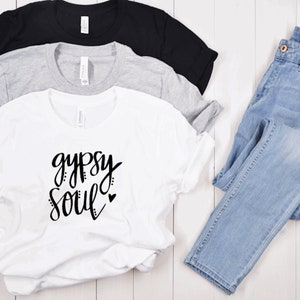 Puede incluir: Camiseta blanca con el texto "gypsy soul" en escritura negra con un peque&ntilde;o coraz&oacute;n. La camiseta est&aacute; doblada y se muestra con otras dos camisetas en negro y gris.