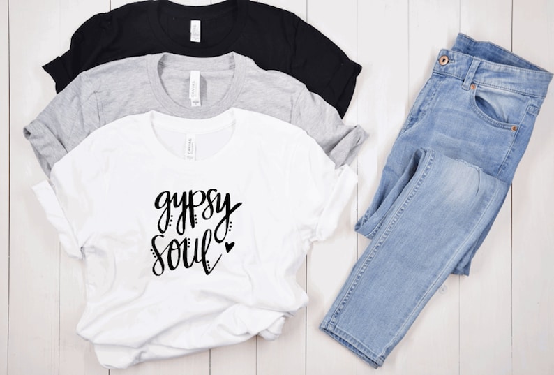 Puede incluir: Camiseta blanca con el texto "gypsy soul" en escritura negra con un peque&ntilde;o coraz&oacute;n. La camiseta est&aacute; doblada y se muestra con otras dos camisetas en negro y gris.