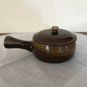 Puede incluir: Un plato de cerámica marrón con tapa y asa. El plato tiene un acabado esmaltado con un esquema de color marrón y beige.