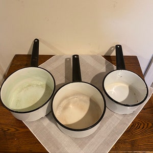 May include: Three white enamel saucepans with black rims and black handles. The saucepans are sitting on a wooden table with a white cloth napkin.