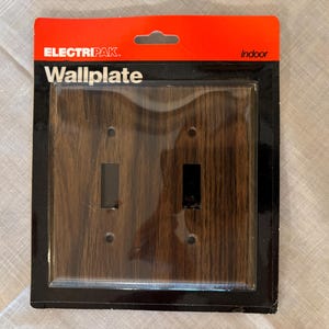 Pode incluir: Uma nova placa de parede para uso interno, com um design de grão de madeira. A placa de parede tem duas aberturas para interruptores e é embalada em plástico transparente. A marca "ELECTRIPAK" e a palavra "Wallplate" estão impressas na embalagem.