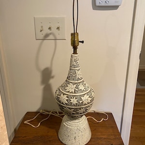 Puede incluir: Una lámpara de mesa de cerámica blanca con un patrón de hojas marrones y blancas. La lámpara tiene una base redonda y una pantalla alta y delgada.