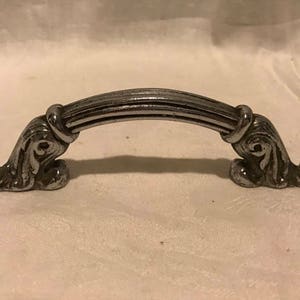 May include: A silver-toned cabinet or drawer pull with a decorative, ornate design. The pull has a curved shape and a smooth, polished finish.