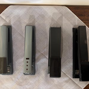 May include: Four staplers of varying colors and designs are arranged on a white patterned cloth. Two are gray, one is black, and one is a darker gray. The staplers are rectangular and have a classic office supply design.