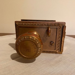 以下が含まれることがあります： 装飾的なデザインと鍵穴のあるアンティーク真鍮製のドアノブ。ノブは装飾的な縁取りのある真鍮プレートに取り付けられています。