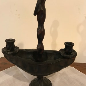 Puede incluir: Un candelero de madera marr&oacute;n oscuro con un dise&ntilde;o &uacute;nico. El candelero tiene dos portavelas y un asa retorcida. La base tiene forma de cuenco con un dise&ntilde;o decorativo.