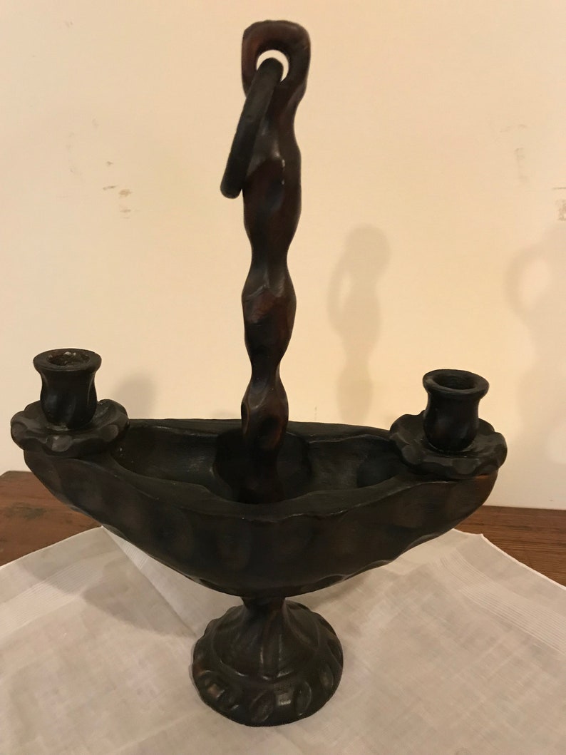 Puede incluir: Un candelero de madera marr&oacute;n oscuro con un dise&ntilde;o &uacute;nico. El candelero tiene dos portavelas y un asa retorcida. La base tiene forma de cuenco con un dise&ntilde;o decorativo.