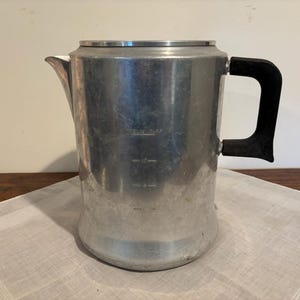May include: A silver-colored aluminum percolator coffee pot with a black handle and a metal lid. The exterior features embossed measurement markings for cup capacity. The body has a cylindrical shape with a tapered base and a pouring spout.