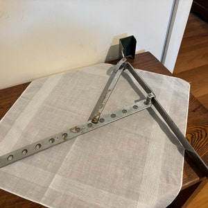May include: A silver metal window stay with a hinge and multiple holes. The stay is in a folded position and is lying on a white cloth.