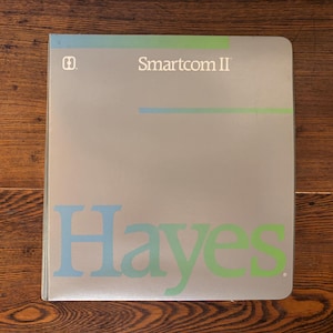 May include: A gray book cover with the title "Smartcom II" and the name "Hayes" in blue and green. The cover has a simple design with horizontal lines in blue and green. The book is on a wooden surface.