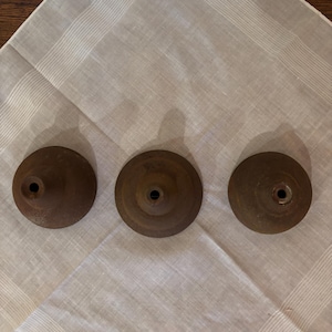 May include: Three antique, brown metal candle holders with a circular base and a central hole. The candle holders are arranged on a white, patterned cloth, creating a vintage aesthetic. The metal has a slightly aged, rustic appearance.